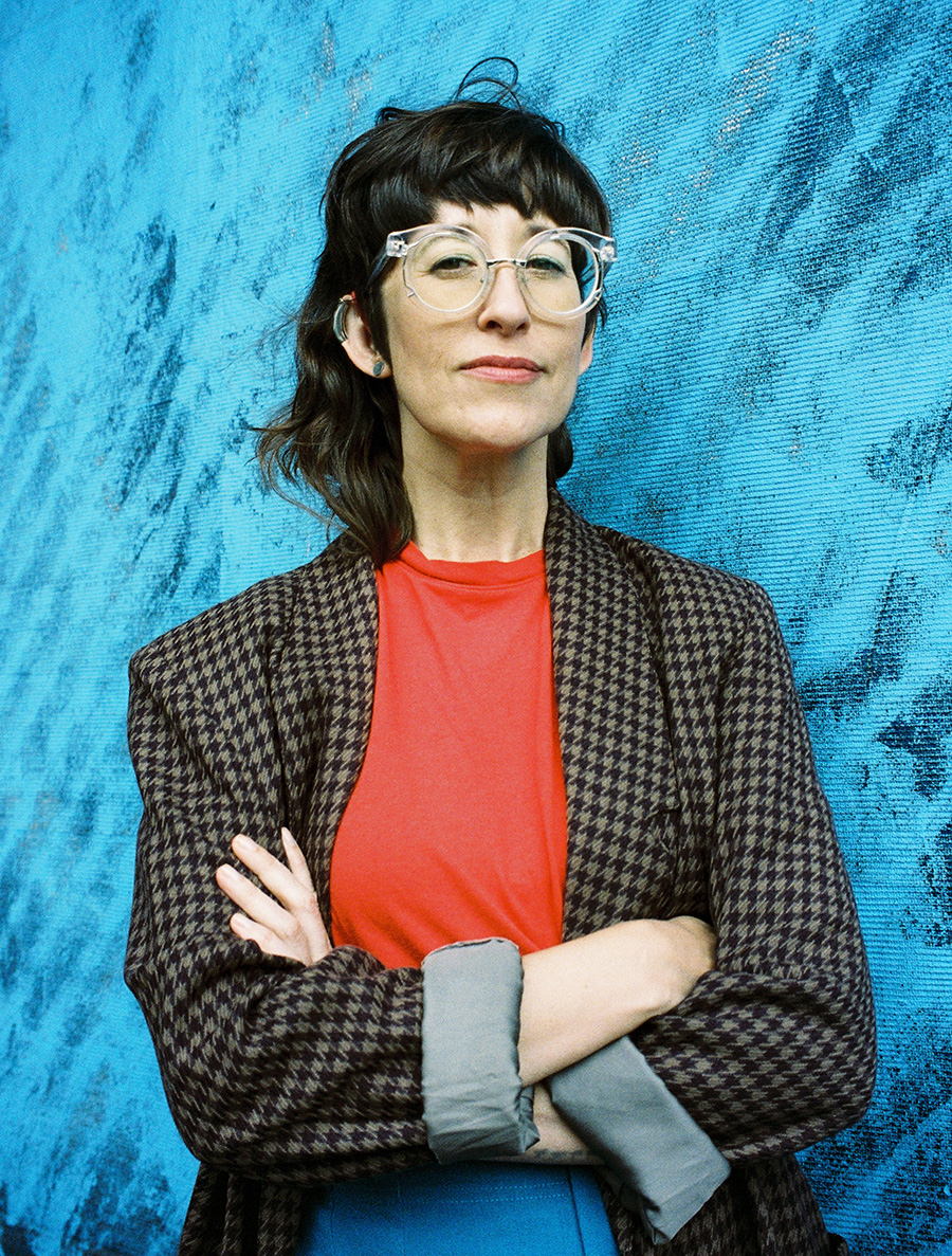 Alison, a white woman with brown bangs and long mullety hair (proudly cut so that her ears and hearing aids are visible) stands with her arms crossed and has a slight smile. She wears bright red, blue and brown clothes and stands in front of a bright blue painted wall.