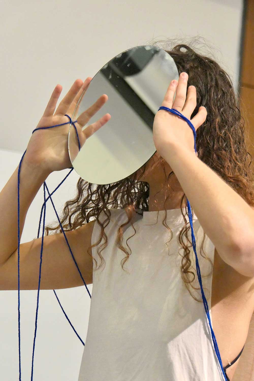 a student holds a round mirror in front of their face while gazing upward, arms dangling thin blue threads