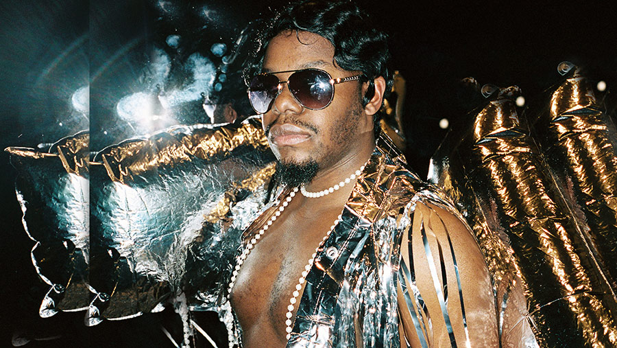 A black man dressed in silver open necked outfit wearing sunglasses surrounded by Mylar balloons