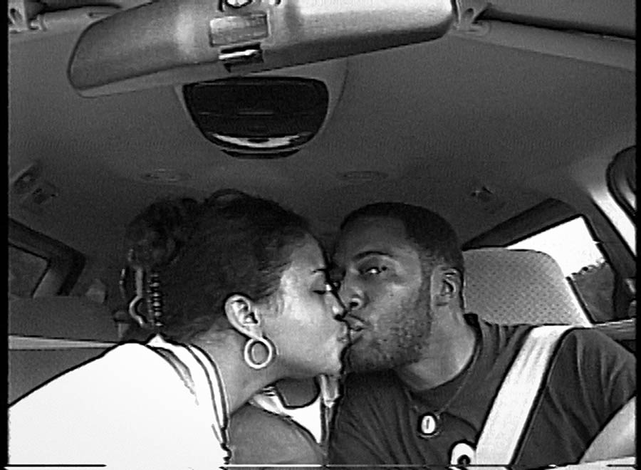 a man and woman kissing in the front seat of a car while the man looks to the camera