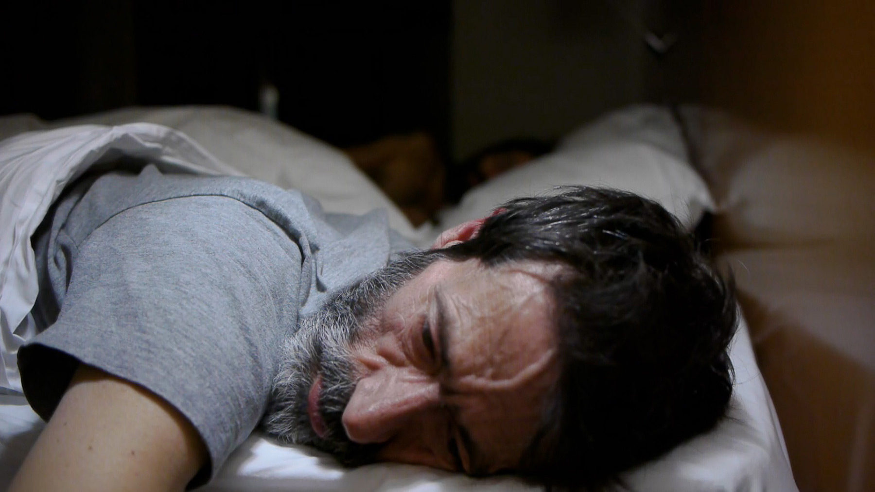 A bearded man lying on his stomach in bed appearing distressed