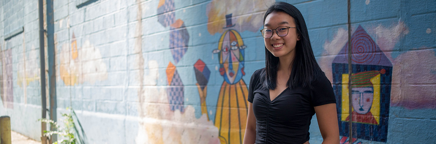 a student poses in front of the side of a building covered in a mural