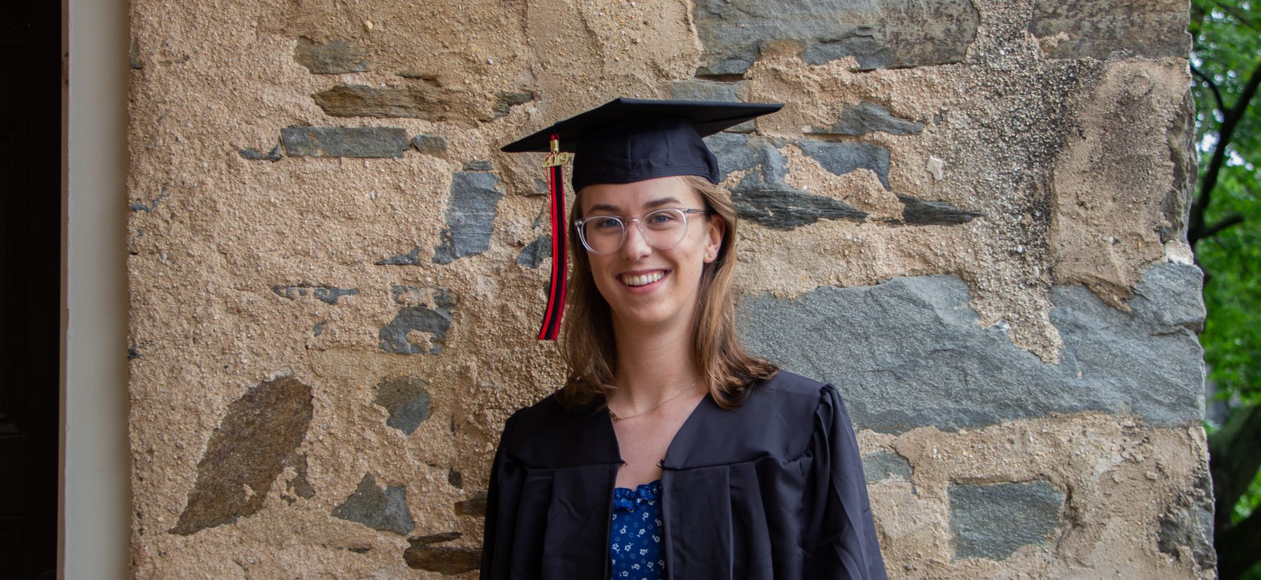 Woman in graduation gown smiles in front of stone wall