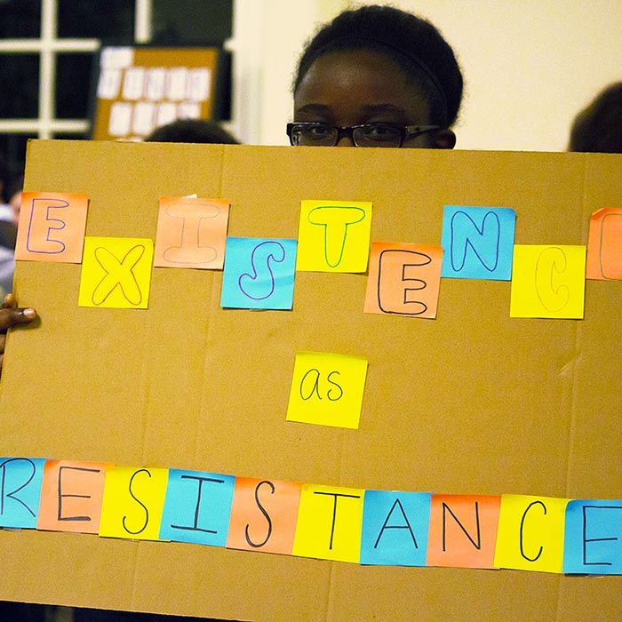 student holds a sign saying Existence as Resistance