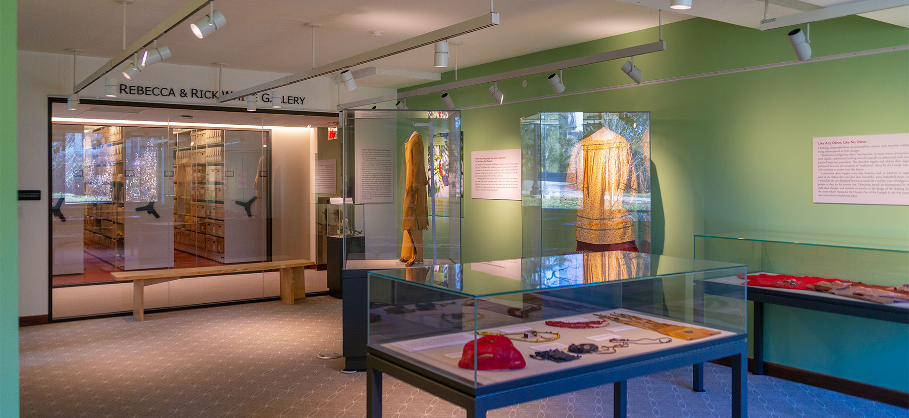 Exhibit space with glass cases displaying clothing. The back wall is green and lit form overhead