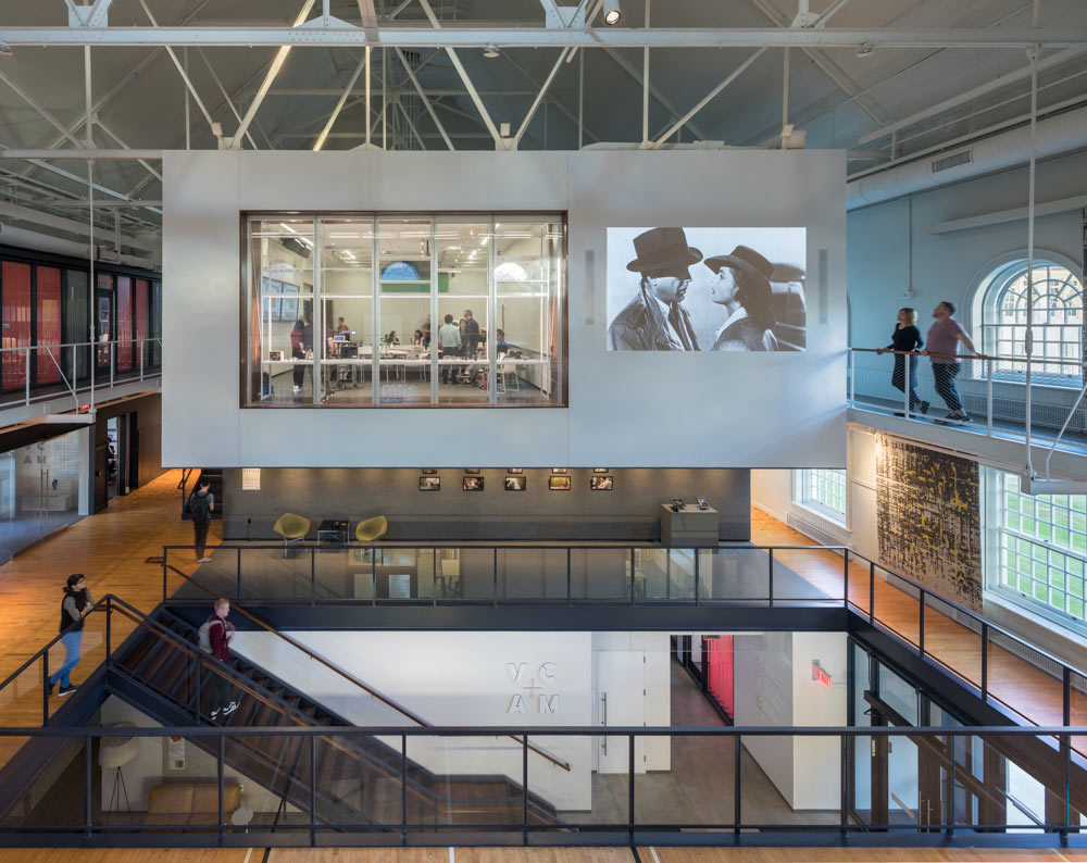 A view into VCAM with a film projected on a wall and people on all three levels