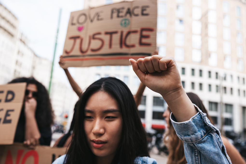 People at a protest. One is holding a sign that reads Peace, Love, Justice.