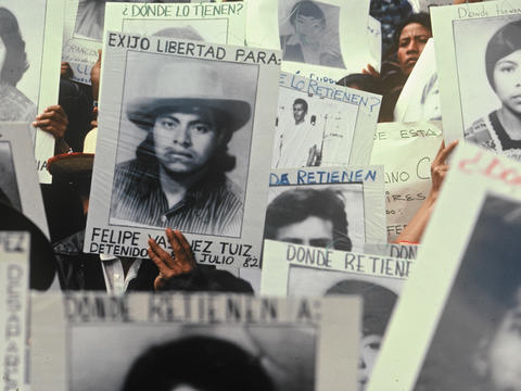 Photo of a crowd of people, mostly only visible by their hands or hats, holding up photos of disappeared loved ones.