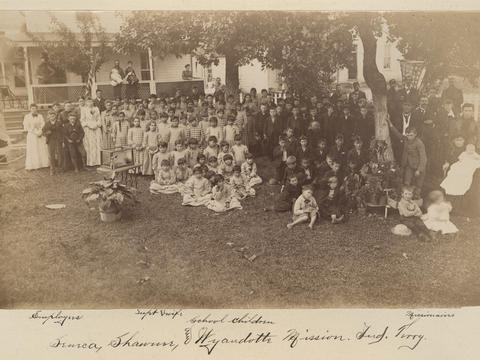 sepia photograph of a crowd of children seemingly separated by gender and a small number of adults, outside in the yard in front of a large house.