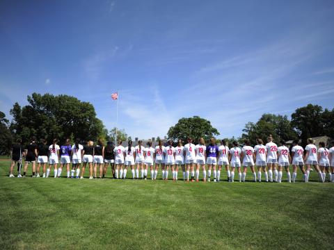 The women's soccer team stands in a line in their uniforms with the American flag waving in the background