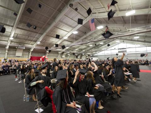 Graduates in black gowns throw their caps in the air inside the Field House at Commencement