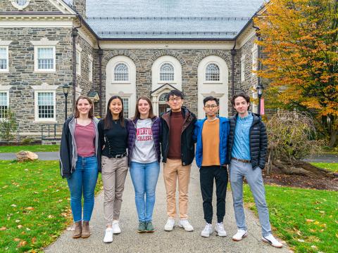 Six students in jackets smile in front of the VCAM building on an autumn day