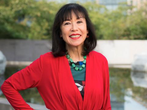 Karen Korematsu smiles in front of a fountain wearing a red jacket