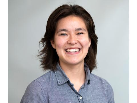 A headshot of Eva Shin smiling in a collared shirt against a gray background