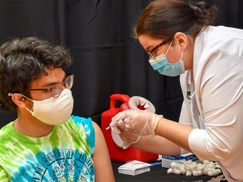A student, wearing a mask and a tie-dye t-shirt, gets the first dose of the Pfizer COVID vaccine in the GIAC