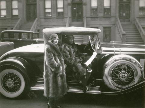 Black and white photograph of a woman in a long fur coat standing next to a man sitting in a classic car. He is also wearing a long fur coat.