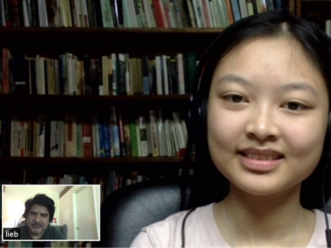 A screen shot of zoom call with a student in a light pink shirt with books in the background. In the corner, there is the view of a man.