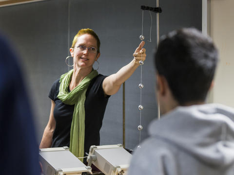 Professor Andrea Lommen, wearing a black shirt, green scarf, and glasses points to a series of metal spheres in a classroom setting.