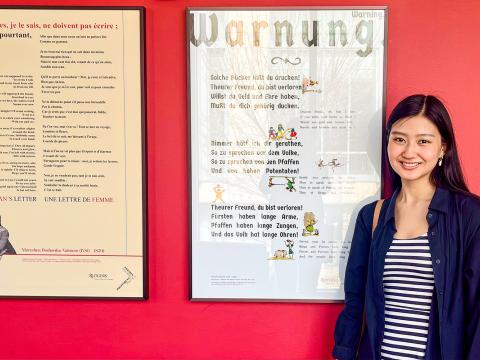 Claire Cai smiles next to a print of Heinrich Heine's poem "Warnung"
