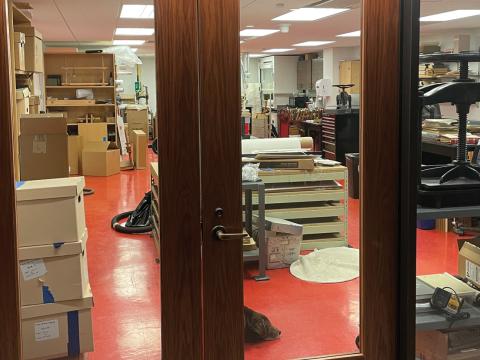 View of the Conservation Lab through the doors and window, a large room filled with shelving and machinery. On the right two people are working at a computer, and a brown dog is asleep on the floor. A sign on the left reads "107 Conservation Lab."