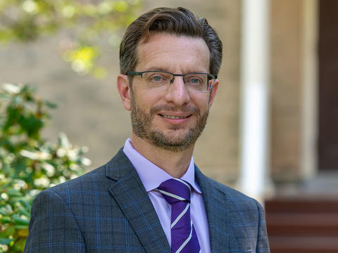 Professor Bret Mulligan wears a grey and blue suit with a lavender shirt and purple tie while standing against and out of focus background