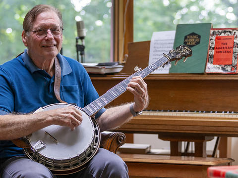 George Stavis holds a banjo and sits on a piano bench in front of a light brown piano.