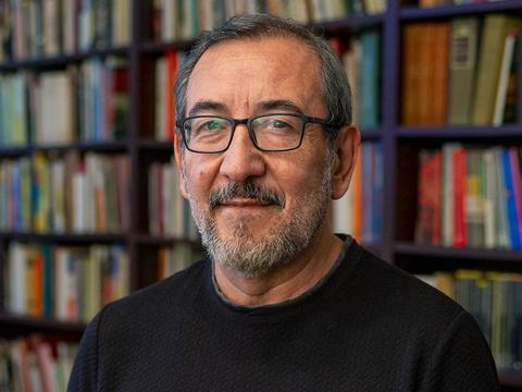 Professor Roberto Castillo Sandoval wears a black shirt and glasses and stands in front of a full bookshelf