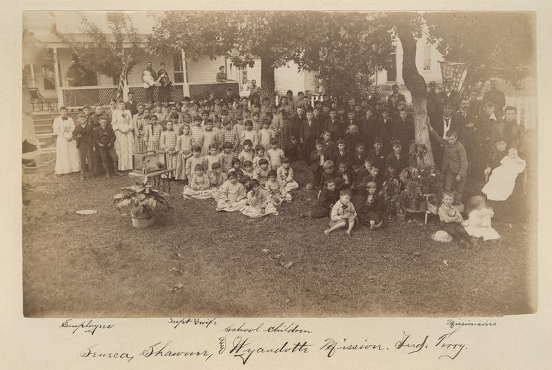 sepia photograph of a crowd of children seemingly separated by gender and a small number of adults, outside in the yard in front of a large house.
