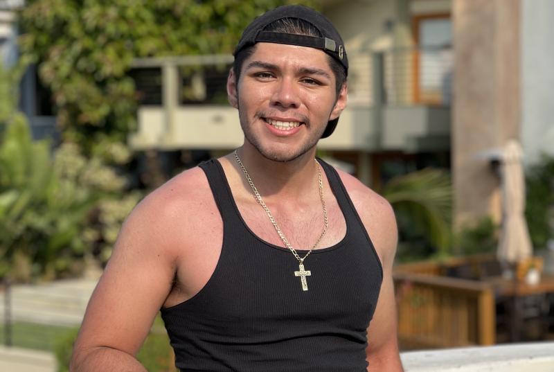 Andrew wears a black tank top, black cap, and cross necklace, and stands in front of a house
