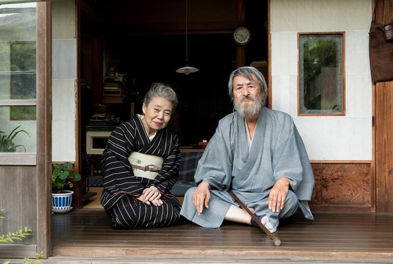 An elderly couple wearing traditional Japanese clothing sit together on a porch