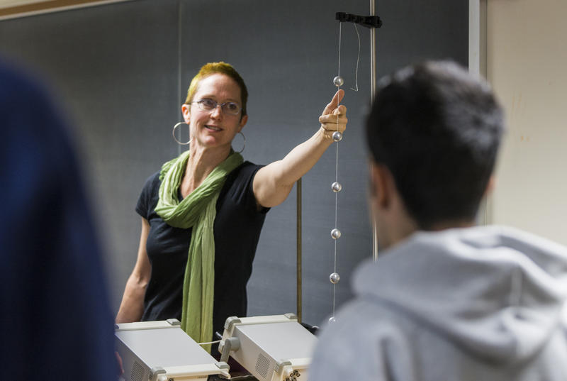 Professor Andrea Lommen, wearing a black shirt, green scarf, and glasses points to a series of metal spheres in a classroom setting.