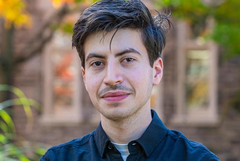 In a portrait, Ahmed Haj Ahmed wears a dark blue collared shirt and stands against a background of an out-of-focus building on Haverford's campus.