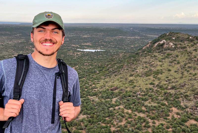 Charlie Mamlin '23 smiles as he stands on a hill overlooking a flat, grassy area in Kenya.