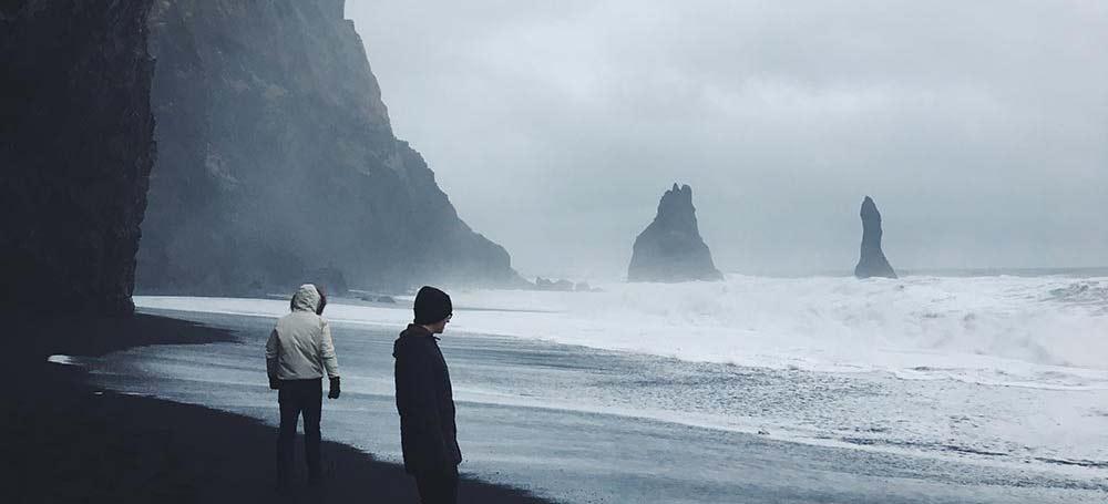 Two figures bundled in winter coats wander a black sand beach with the ocean roaring in the background