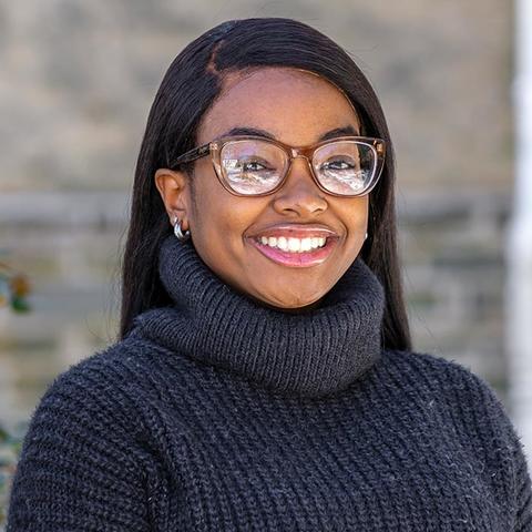 Maye-Gan Brown in a black knitted sweater and glasses, smiling for the camera