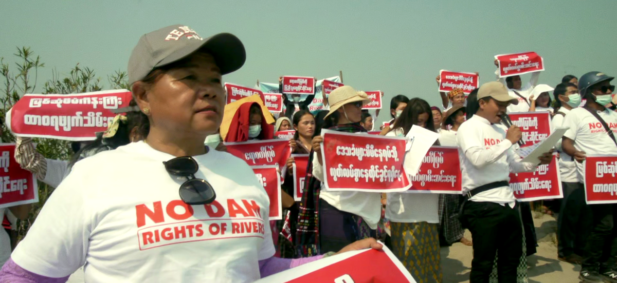 A film still from Emily Hong's "Above and Below the Ground" portrays protestors in Myanmar wearing white shirts and carrying red protest signs with white lettering.