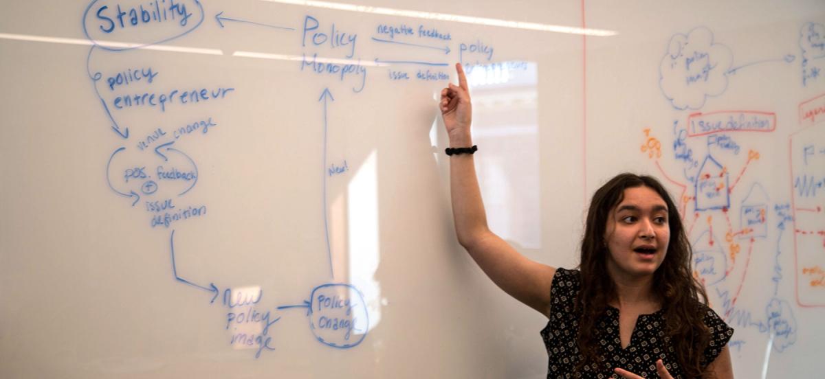 Lilian Feist stands in front of a whiteboard covered with writing while explaining her diagram