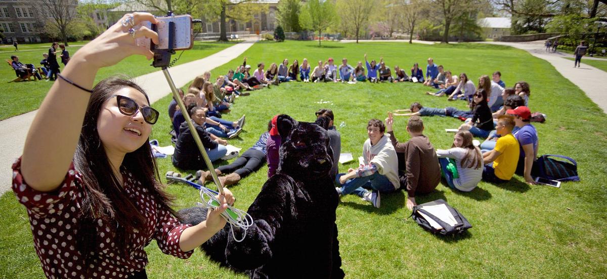 Black squirrel mascot taking a selfie with students sitting in a circle on grass.