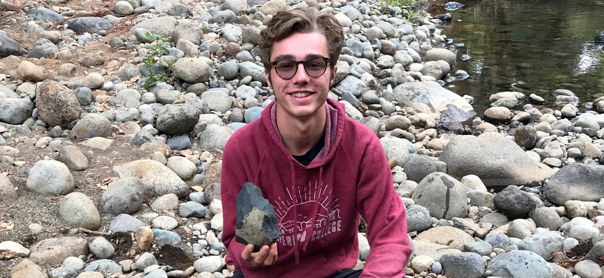 Levi crouches in a field of stones holding a handaxe.