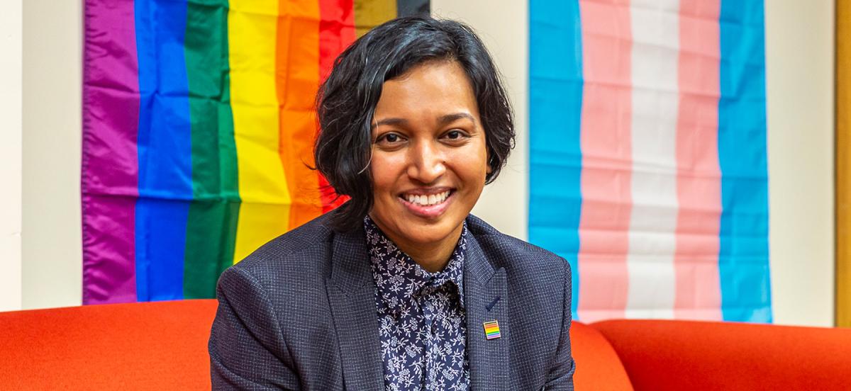 Sayeeda smiles, sitting in the GRASE office in front of a Pride flag and a transgender flag