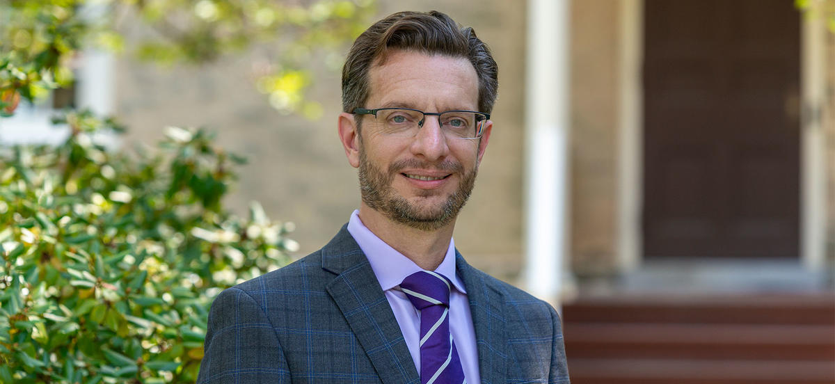 Professor Bret Mulligan wears a grey and blue suit with a lavender shirt and purple tie while standing against and out of focus background