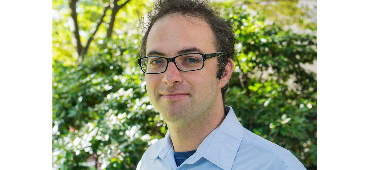 Professor Daniel Grin stands in front of greenery wearing a blue button down shirt.