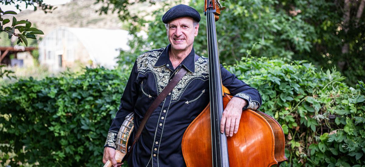 Mark Schatz '78 holds his string bass and his banjo in front of a background of bushes. Photo by Lisa Berman.
