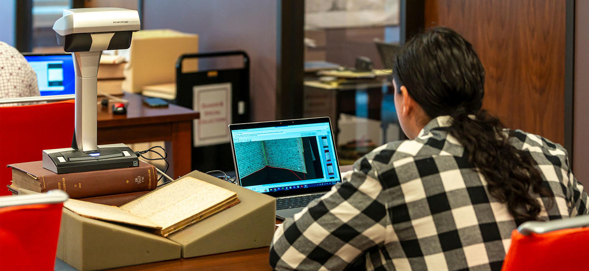 A man in a black and white checkered shirt uses an overhead scanner to process documents related to Quaker run Native American Boarding Schools.