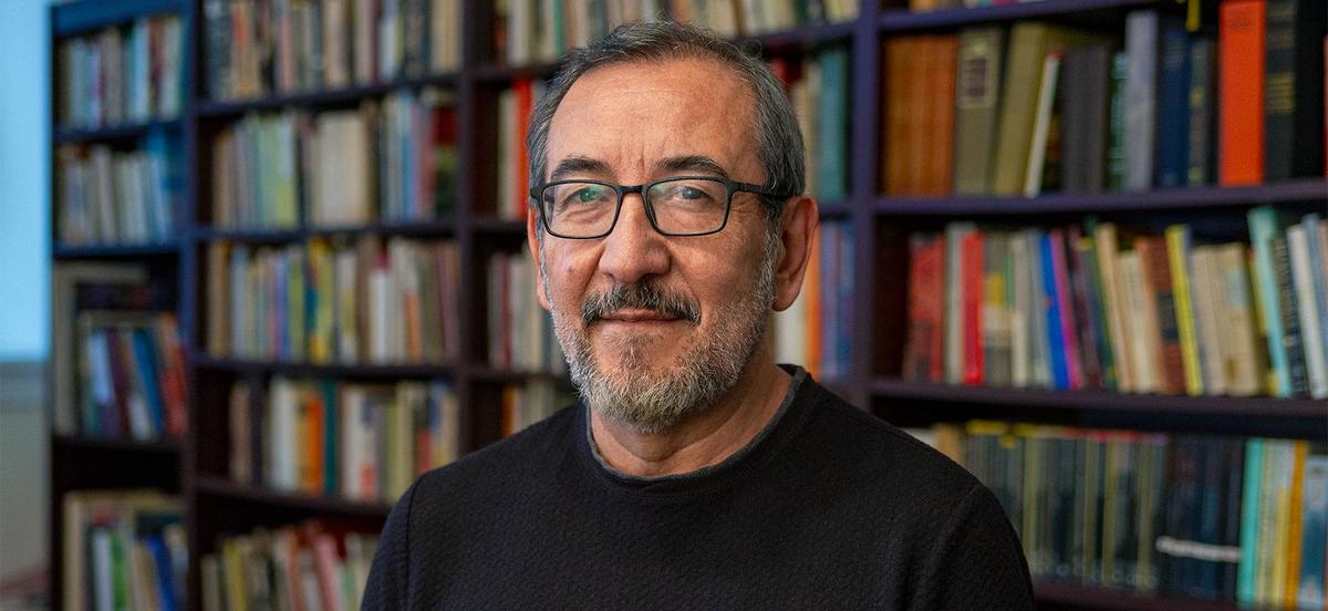 Professor Roberto Castillo Sandoval wears a black shirt and glasses and stands in front of a full bookshelf